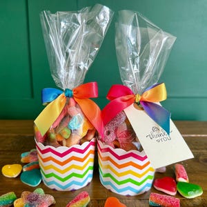May include: Two clear plastic bags filled with assorted colourful gummy sweets, each sitting in a white paper cup with a vibrant rainbow chevron pattern. Each bag is tied with a multi-coloured satin ribbon and one features a tag with the text thank you.