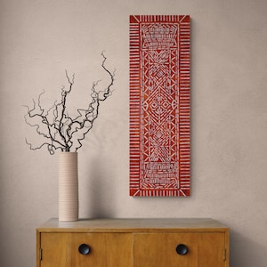 May include: A tall, rectangular wall art piece featuring a vibrant red background with intricate, repeating white geometric patterns and lines. The artwork is displayed vertically, showcasing a symmetrical design of dashes and shapes across the surface.