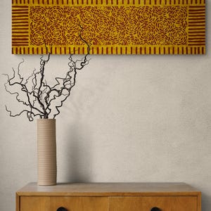 May include: A rectangular wall art piece featuring a mustard yellow background with a repeating pattern of dark red concentric circles and vertical stripes along the borders. The artwork is displayed above a wooden cabinet with two black circular knobs.