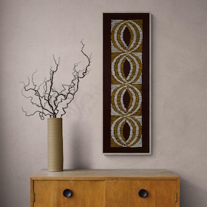 May include: A brown wooden dresser with two drawers and gold knobs. A vase with a dried branch arrangement sits on top of the dresser. A framed artwork with a geometric pattern in gold and white hangs on the wall behind the dresser.