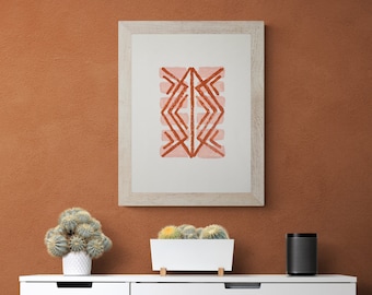 Puede incluir: Una imagen enmarcada en blanco con un diseño geométrico en tonos rosa y naranja cuelga de una pared de terracota. Una pequeña planta de cactus en maceta se encuentra sobre una cómoda blanca con un altavoz negro.