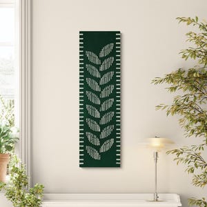 May include: A dark green rectangular wall art piece with a white leaf design. The artwork has a white border with vertical lines. The piece is mounted on a cream-colored wall, with a lamp and greenery in the background.