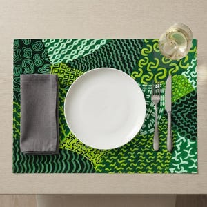 May include: A rectangular placemat featuring a patchwork of various green and black geometric patterns. A solid white plate sits in the center, accompanied by a grey cloth napkin on the left and a silver fork and knife on the right side.