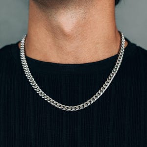 Peut inclure: Un collier chaîne gourmette argenté. Le collier est composé de maillons entrelacés et est porté sur un t-shirt noir côtelé. L'arrière-plan est de couleur grise unie.