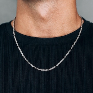 May include: A silver chain necklace with a circular link design. The necklace is worn against a black ribbed top. The background is a solid grey colour.