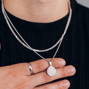 May include: A silver chain necklace with a round pendant featuring a detailed design. The necklace is layered over a second, thinner silver chain.