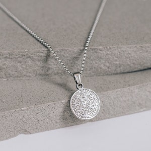 May include: A silver chain necklace with a round pendant featuring a detailed design. The pendant is on a grey textured surface.