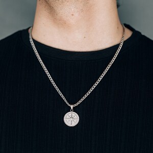 May include: A silver chain necklace with a round pendant featuring a detailed design. The necklace is worn by a person with a black shirt.