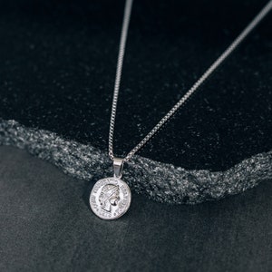 May include: A silver chain necklace with a silver coin pendant featuring a profile of a woman's face. The necklace is on a dark gray textured surface.