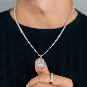 May include: Silver-toned necklace with a rectangular pendant featuring a sunburst design. The pendant hangs from a chain. A matching silver ring is also visible. The person is wearing a black shirt.