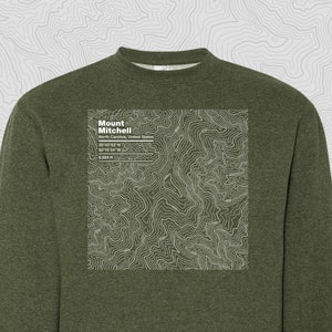May include: A green sweatshirt with a white topographic map design featuring Mount Mitchell, North Carolina, United States. The map includes the coordinates 35°45′53″N, 82°15′54″W, and the elevation 6,684 ft.