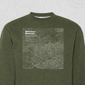 Puede incluir: Una sudadera verde con un estampado gráfico blanco de un mapa topográfico de la montaña Whiteface en Nueva York, Estados Unidos. El gráfico incluye el nombre de la montaña, su ubicación, coordenadas y elevación: 4.867 pies.