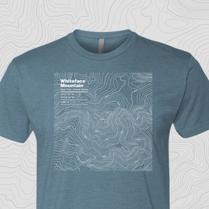 Puede incluir: Una camiseta azul con un diseño de mapa topográfico blanco de la montaña Whiteface en Nueva York, Estados Unidos. El mapa incluye las coordenadas 44°21'57"N 73°54'10"W y la elevación 4.867 pies.