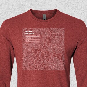 May include: A maroon long-sleeve shirt featuring a white topographic map of Mount Mitchell, North Carolina. The map includes the mountain's coordinates and elevation of 6,684 ft. The text "Mount Mitchell" is displayed at the top of the map.