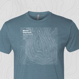 May include: A teal blue t-shirt with a white topographic map design of Mount Haystack in New York, United States. The map includes the mountain's coordinates and elevation: 44°06′20″N, 73°54'02"W, 4,960 ft.