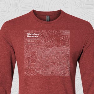 Puede incluir: Una camiseta de manga larga de color rojo ladrillo con un mapa topográfico blanco del monte Whiteface en Nueva York, con coordenadas y detalles de elevación. El diseño incluye el texto "Whiteface Mountain".
