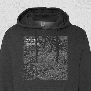 Puede incluir: Sudadera con capucha gris oscuro con un mapa topográfico blanco de Whiteface Mountain, Nueva York, con coordenadas y elevación. La sudadera tiene una capucha con cordón y un bolsillo de canguro.