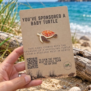 May include: A tan rectangular card featuring an enamel sea turtle pin with an orange and brown shell. The text reads: YOU'VE SPONSORED A BABY TURTLE, THIS CARD COMES WITH THE SPONSOR OF A HATCHLING TURTLE FROM A RESCUED NEST IN KENYA, and a QR code.