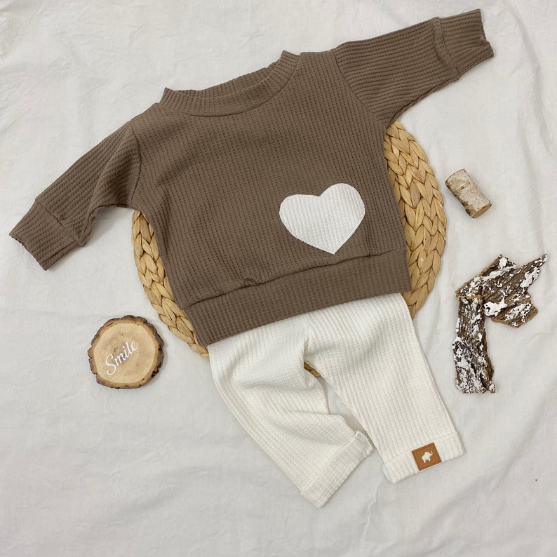 May include: A brown waffle knit long-sleeve shirt with a white heart on the front, paired with white waffle knit pants. The outfit is laid out on a white surface with a wooden sign that says "Smile".