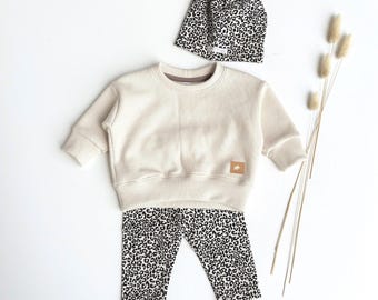 May include: A cream-colored waffle-knit long-sleeve sweatshirt with a small tan label, paired with leopard-print leggings and a matching leopard-print beanie. The set is displayed alongside three dried decorative grass stems on a plain surface.