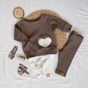 May include: A brown waffle knit baby sweater with a white heart applique and matching brown waffle knit leggings. The sweater has a crew neck and long sleeves. The leggings have a white waffle knit waistband.