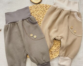 May include: Two pairs of waffle knit baby pants. One pair is brown with brown trim and the other is beige with a beige trim. Both pairs have a pocket on the back with a button.
