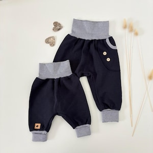 Könnte beinhalten: Zwei marineblaue Babyhosen mit strukturiertem Waffelstrick. Die Hosen haben einen hohen, gestreiften Bund und Bündchen. Eine Hose hat eine kleine Tasche mit zwei Holzknöpfen. Ein kleines braunes Lederetikett befindet sich an einem Hosenbein.