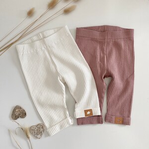 May include: Two pairs of waffle knit leggings, one white and one dusty rose. Both pairs have a small brown patch with a white bear logo on the bottom of each leg.