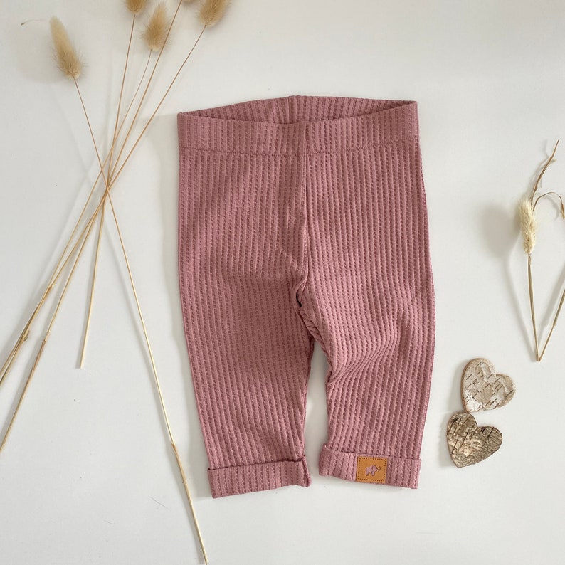 May include: A pair of pink waffle knit leggings with cuffed ankles. The leggings have a small brown patch with a white logo on the right leg.