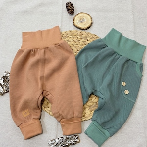 Könnte beinhalten: Zwei Paar Babyhosen, eine in Terrakotta und eine in Teal-Grün. Beide Hosen haben eine Waffelstruktur und einen breiten elastischen Bund. Die Teal-Hose hat ein Knopfdetail an der Gesäßtasche.