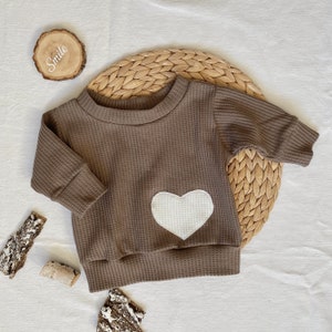May include: A brown waffle knit long-sleeved shirt with a white heart applique. The shirt is on a woven straw mat.
