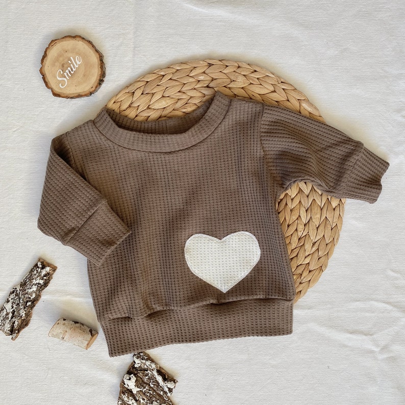May include: A brown waffle knit long-sleeved shirt with a white heart applique. The shirt is on a woven straw mat.
