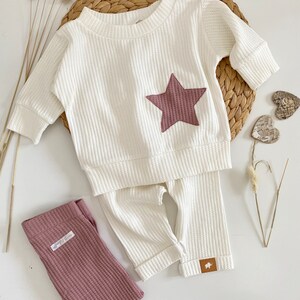 May include: White waffle knit baby outfit with a brown star on the top and matching pants. The pants have a brown label with a small animal on it.