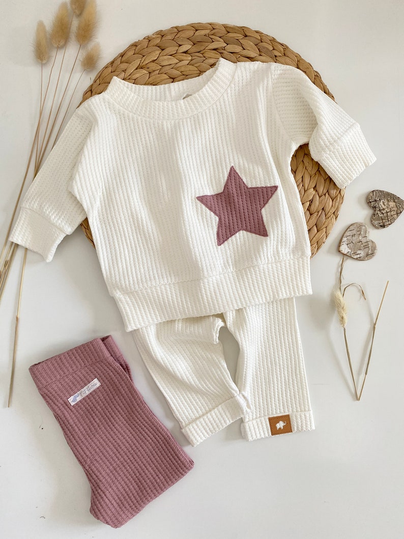 May include: White waffle knit baby outfit with a brown star on the top and matching pants. The pants have a brown label with a small animal on it.