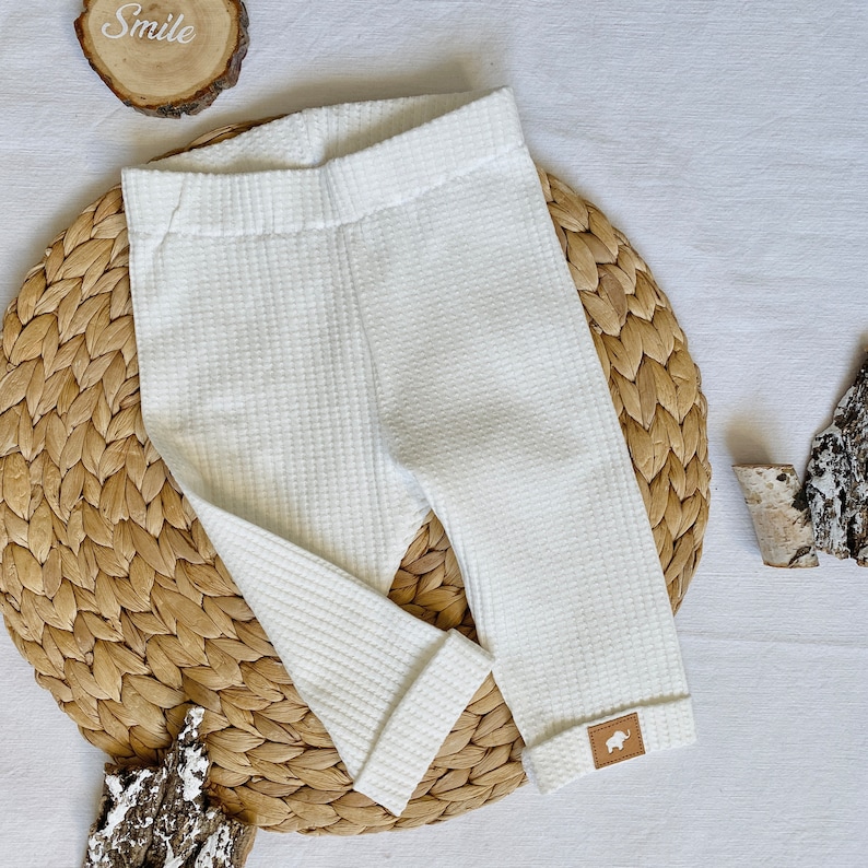 May include: White waffle knit leggings with a small brown leather patch on the leg. The leggings are on a woven straw mat.