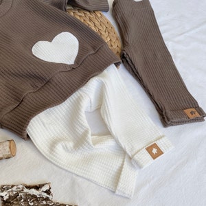 May include: A brown waffle knit sweater with a white heart on the front and matching brown waffle knit leggings. The leggings have a small brown patch with a white animal silhouette on the bottom of the leg.