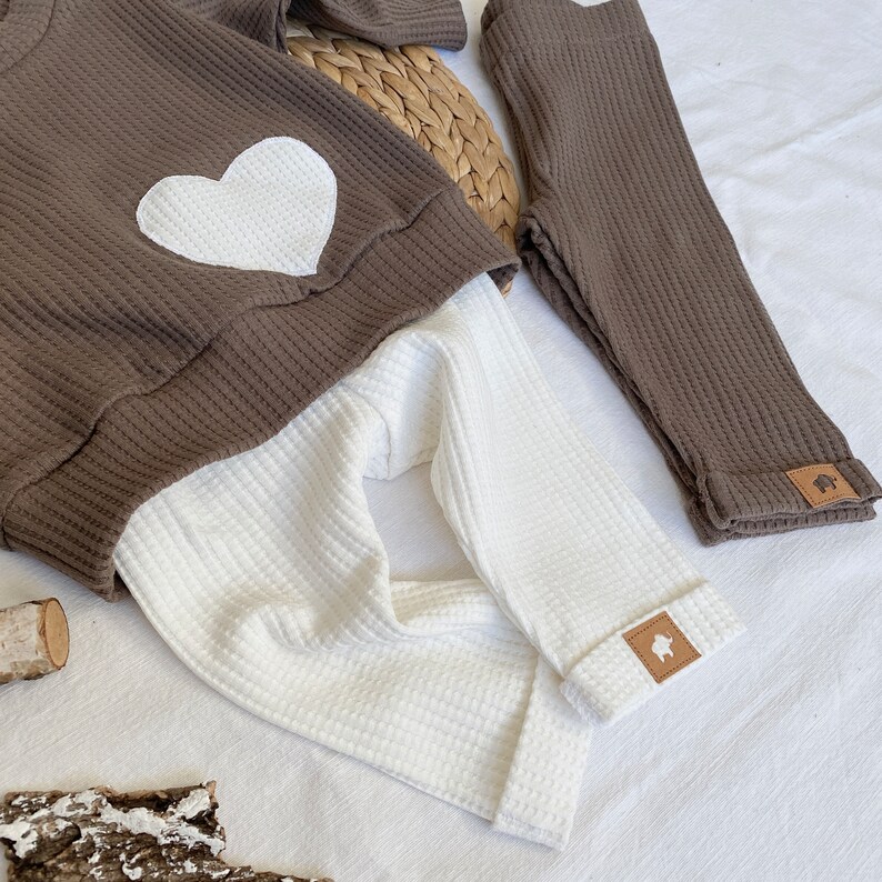 May include: A brown waffle knit sweater with a white heart on the front and matching brown waffle knit leggings. The leggings have a small brown patch with a white animal silhouette on the bottom of the leg.
