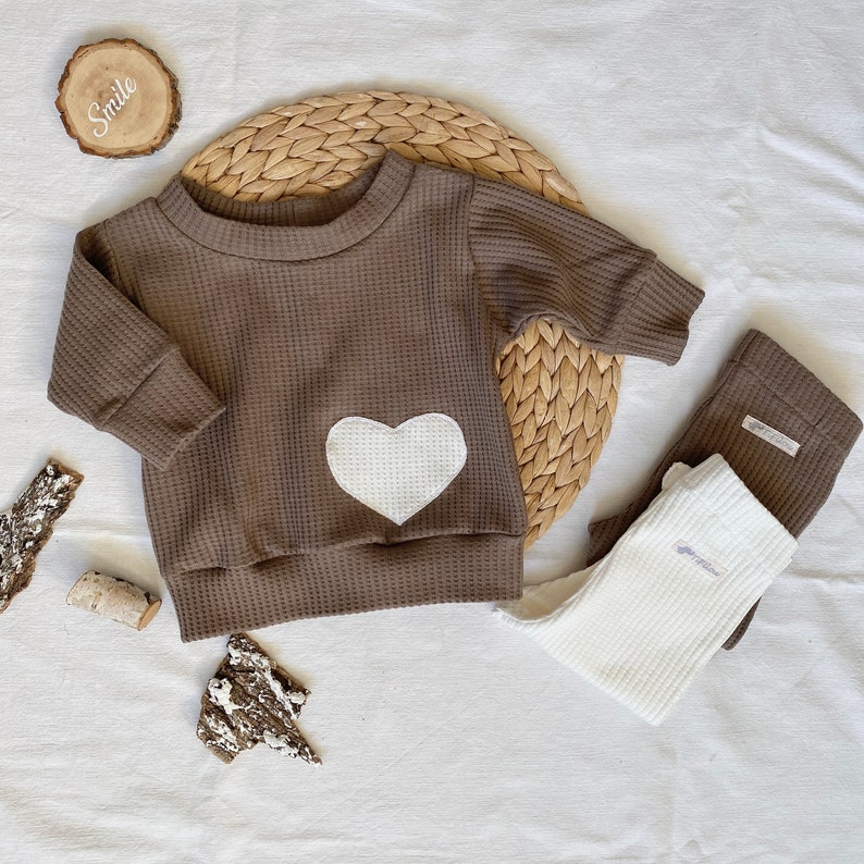 May include: A brown waffle knit long-sleeved shirt with a white heart-shaped patch on the front. The shirt is paired with white waffle knit leggings. The image also includes a wooden sign with the word "Smile" on it.