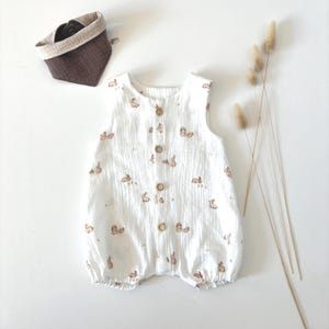 Könnte beinhalten: Weißer Baby-Strampler mit einem Muster aus braunen Hasen. Der ärmellose Strampler hat Holzknöpfe vorne und einen gerafften Saum. Ein braunes Lätzchen und getrocknetes Gras sind ebenfalls im Bild.