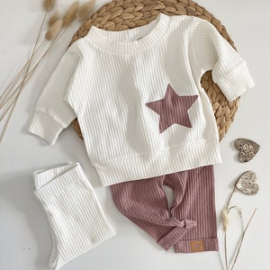 May include: A white waffle knit baby onesie with a pink star on the front and matching pink waffle knit pants. The pants have a knotted detail at the bottom.