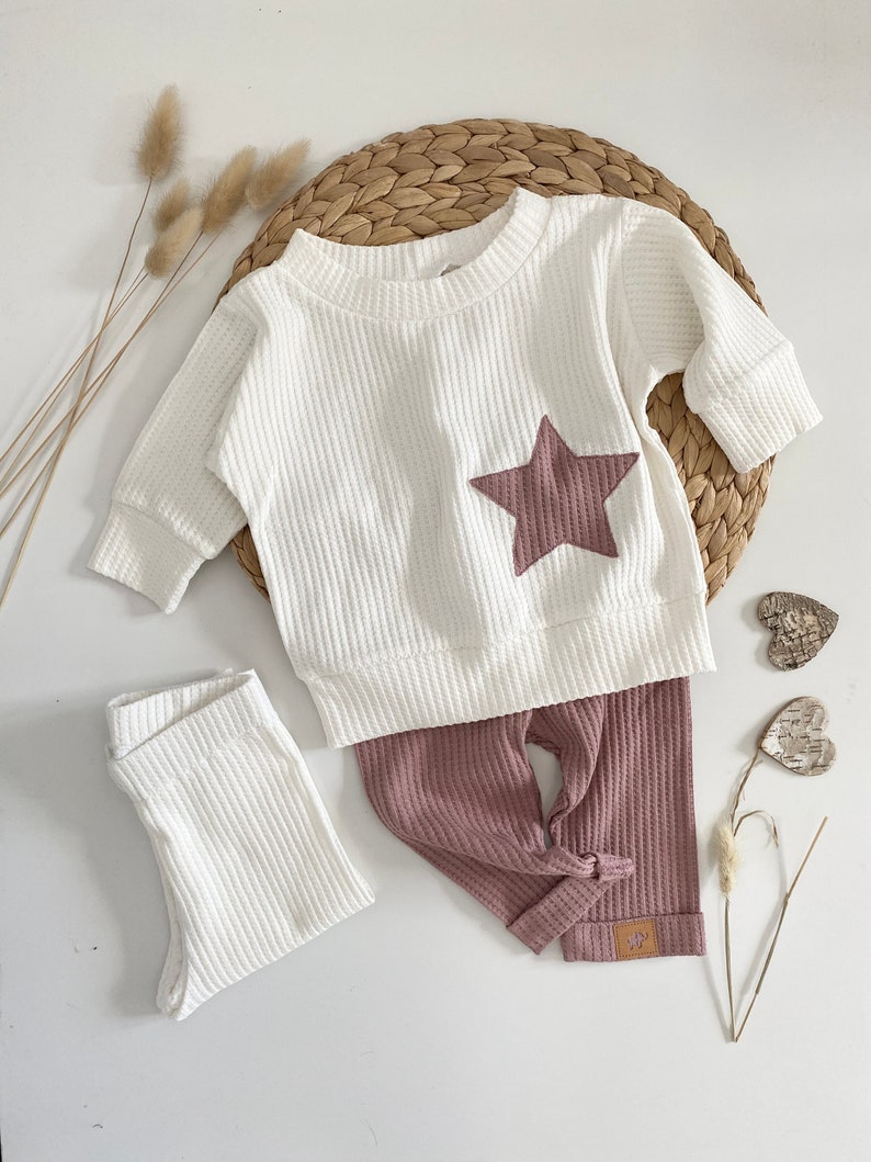 May include: A white waffle knit baby onesie with a pink star on the front and matching pink waffle knit pants. The pants have a knotted detail at the bottom.