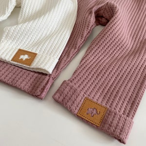 May include: Two pairs of waffle knit pants with brown leather patches featuring an elephant silhouette. One pair is white and the other is mauve.
