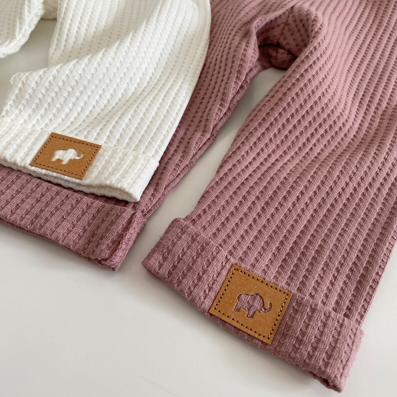 May include: Two pairs of waffle knit pants with brown leather patches featuring an elephant silhouette. One pair is white and the other is mauve.