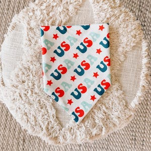 May include: White bandana with red, blue, and light blue text and red star graphics. The bandana features the word "USA" repeated multiple times. The bandana is a patriotic accessory.