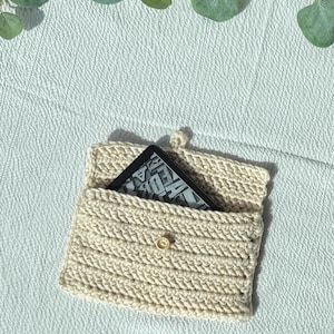 Puede incluir: Una funda de lector electrónico de ganchillo de color crema con cierre de botón. La funda contiene un lector electrónico negro con texto visible en la pantalla. La funda está hecha de hilo grueso y texturizado y tiene una solapa.