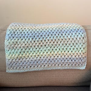 以下が含まれることがあります： 青、緑、クリームの柔らかなパステルカラーの手作りのかぎ針編みブランケット。ブランケットは、触覚的な表面を作り出す、テクスチャード加工された隆起したステッチパターンを備えています。ブランケットはニュートラルなトーンのソファに掛けられています。