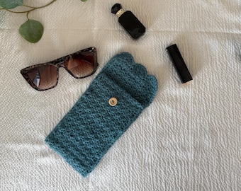 May include: Teal crochet sunglasses case with a scalloped top and a gold button closure. Tortoiseshell sunglasses, a black perfume bottle, and a tube of lipstick are also visible.