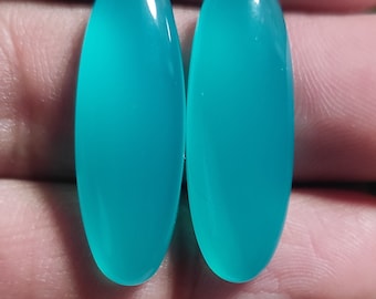 Könnte beinhalten: Zwei ovale türkisfarbene Cabochons, jeder mit einer glatten, polierten Oberfläche. Die Cabochons sind in einem leuchtenden Blaugrün gehalten und haben eine durchscheinende Optik.