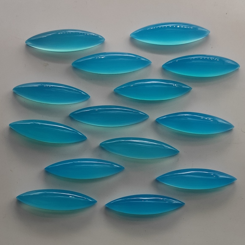 Puede incluir: Una colecci&oacute;n de 13 piedras preciosas de turquesa en cabuj&oacute;n, cada una con forma ovalada y un extremo puntiagudo. Las piedras est&aacute;n dispuestas en forma escalonada sobre una superficie blanca.