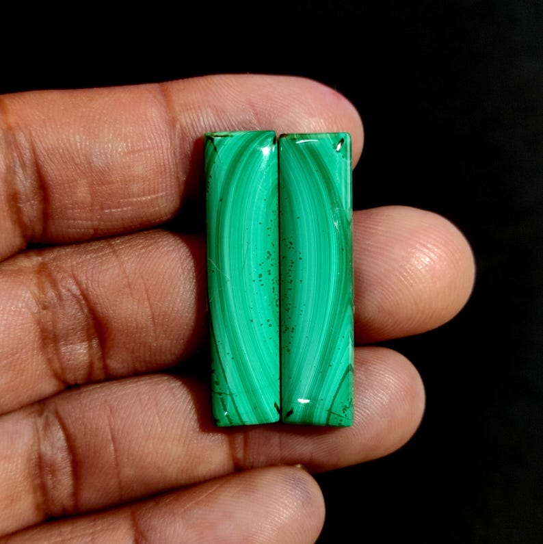 May include: Two rectangular malachite stones with a vibrant green color and swirling patterns. The polished surfaces reflect light, showcasing the natural beauty of the mineral. The stones are held in a hand against a dark background.